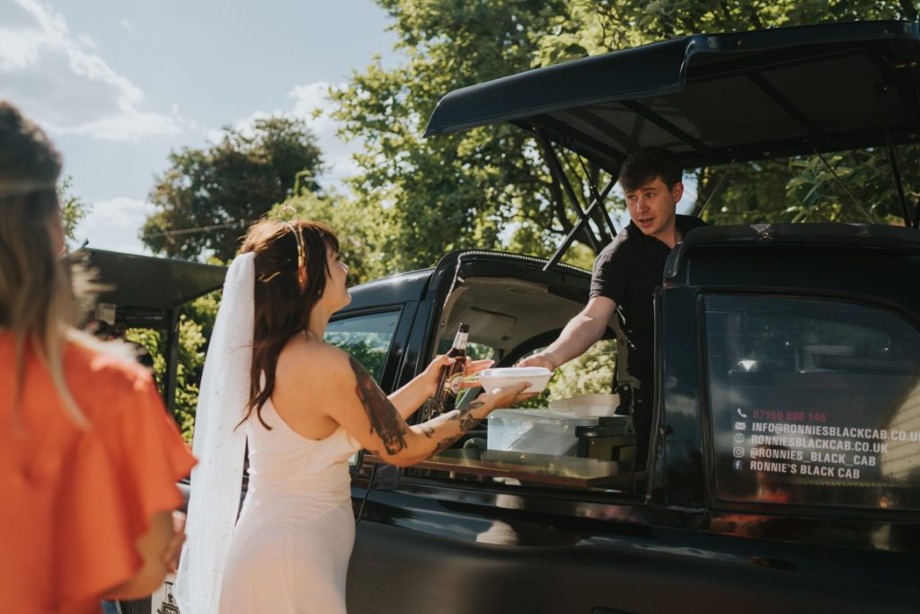 A man handing a bride a pie out of his black cab which is a street food van.